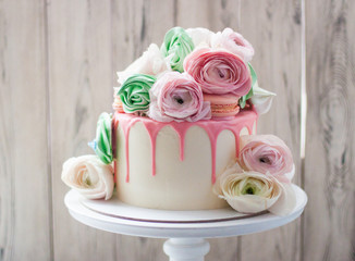 White cake with pink melted chocolate decorated with fresh flowers, roses, ranunculuses, macaroons and meringue swirls. 