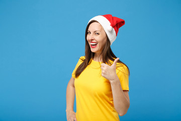 Laughing young brunette woman Santa girl in yellow t-shirt Christmas hat posing isolated on blue wall background. Happy New Year 2020 celebration holiday concept. Mock up copy space. Showing thumb up.