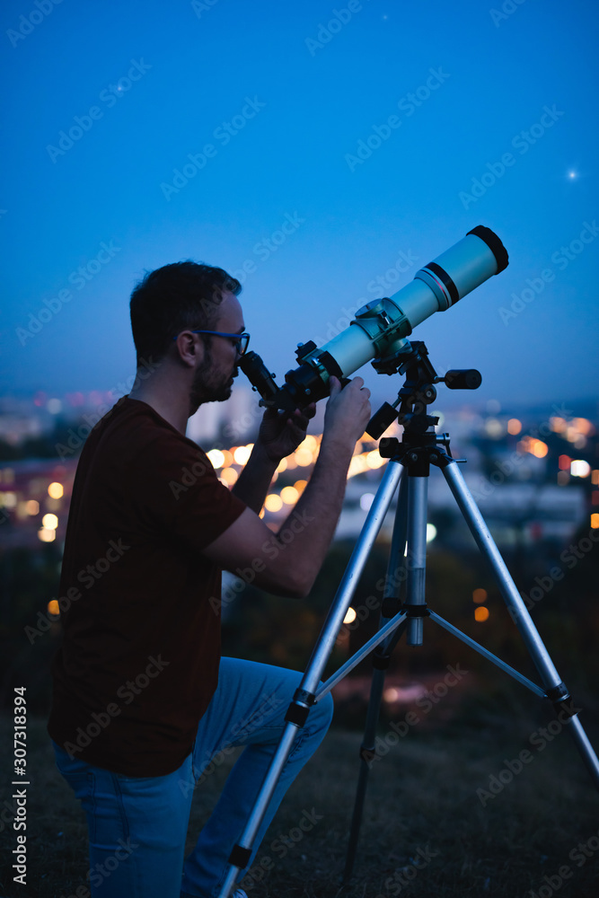 Wall mural astronomer with a telescope watching at the stars and moon with blurred city lights in the backgroun
