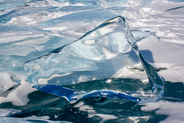 A large ice floe melted by the sun stands on the ice of Lake Baikal. Reflections and distortions in transparent ice. Snow is around. Green and blue color of ice.