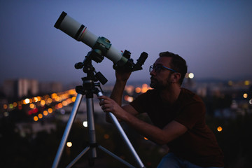 Astronomer with a telescope watching at the stars and Moon with blurred city lights in the...