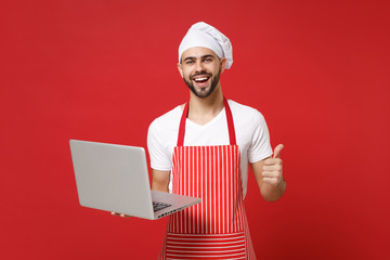Laughing young bearded chef cook or baker man in striped apron t-shirt toque chefs hat isolated on red background. Cooking food concept. Mock up copy space. Hold laptop pc computer, showing thumb up.