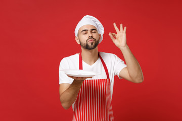 Chef cook or baker man in striped apron toque chefs hat isolated on red background. Cooking food concept. Mock up copy space. Hold empty blank plate with place for food making okay taste delight sign.