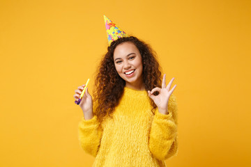 Smiling young african american girl in fur sweater, birthday hat posing isolated on yellow orange background in studio. People lifestyle concept. Mock up copy space. Holding pipe, showing OK gesture.