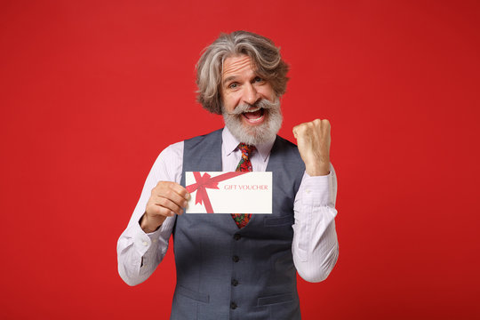 Joyful elderly gray-haired mustache bearded man in classic shirt vest colorful tie isolated on red background. People lifestyle concept. Mock up copy space. Hold gift certificate doing winner gesture. - Powered by Adobe