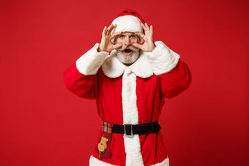 Fototapeta na wymiar Shocked elderly gray-haired mustache bearded Santa man in Christmas hat posing isolated on red wall background. Happy New Year 2020 celebration holiday concept. Mock up copy space. Stretching eyelids.