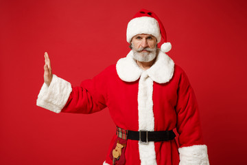 Elderly gray-haired bearded mustache Santa man in Christmas hat posing isolated on red background. New Year 2020 celebration holiday concept. Mock up copy space. Showing stop gesture aside with palm.