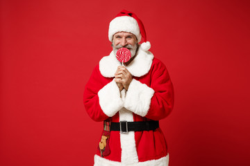 Fototapeta na wymiar Smiling Elderly gray-haired mustache bearded Santa man in Christmas hat posing isolated on red background. Happy New Year 2020 celebration holiday concept. Mock up copy space. Holding round lollipop.