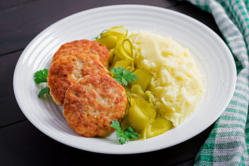 Homemade fried cutlets/meatballs with mashed potatoes and pickled cucumber on white plate.