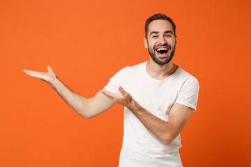 Laughing funny young man in casual white t-shirt posing isolated on bright orange wall background, studio portrait. People sincere emotions lifestyle concept. Mock up copy space. Pointing hands aside.