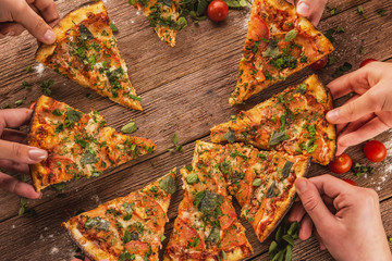 Hands reach for pizza, which lies on a wooden table with cherry tomatoes