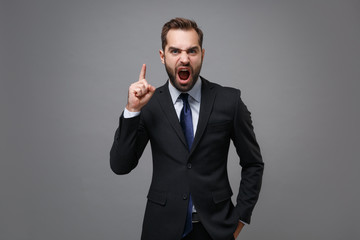 Angry young business man in classic black suit shirt tie posing isolated on grey background. Achievement career wealth business concept. Mock up copy space. Point index finger up, screaming swearing.