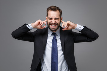 Frustrated young business man in suit shirt tie posing isolated on grey background. Achievement career wealth business concept. Mock up copy space. Covering ears with fingers, keeping eyes closed.