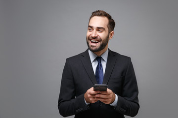 Laughing young business man in suit shirt tie posing isolated on grey background. Achievement career wealth business concept. Mock up copy space. Using mobile phone typing sms message, looking aside.