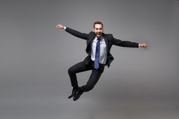 Cheerful young business man in classic black suit shirt tie posing isolated on grey background. Achievement career wealth business concept. Mock up copy space. Jumping, spreading hands, having fun.