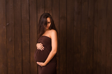 Fototapeta premium Pregnant woman in brown dress holds hands on belly on a dark brown background. Pregnancy, maternity, expectation concept. Beautiful tender mood photo of pregnancy