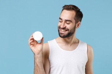 Bearded young man 20s years old applying moisturizer facial cream from container isolated on blue...