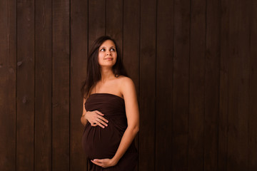Fototapeta premium Pregnant woman in brown dress holds hands on belly on a dark brown background. Pregnancy, maternity, expectation concept. Beautiful tender mood photo of pregnancy