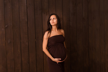 Fototapeta premium Pregnant woman in brown dress holds hands on belly on a dark brown background. Pregnancy, maternity, expectation concept. Beautiful tender mood photo of pregnancy