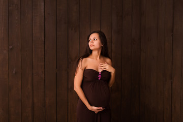 Fototapeta premium Pregnant woman in brown dress holds hands on belly on a dark brown background. Pregnancy, maternity, expectation concept. Beautiful tender mood photo of pregnancy