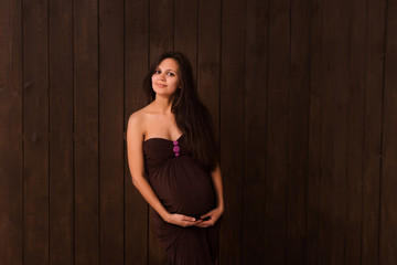 Pregnant woman in brown dress holds hands on belly on a dark brown background. Pregnancy, maternity, expectation concept. Beautiful tender mood photo of pregnancy