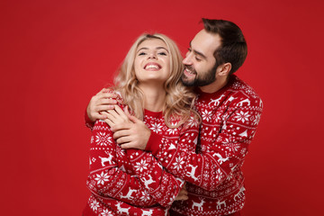 Funny young couple guy girl in Christmas knitted sweaters posing isolated on bright red background studio portrait. Happy New Year 2020 celebration holiday party concept. Mock up copy space. Hugging.
