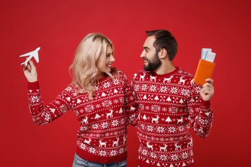 Smiling young couple guy girl in Christmas sweaters posing isolated on red background. Happy New Year 2020 celebration holiday concept. Mock up copy space. Hold passport boarding pass ticket airplane.