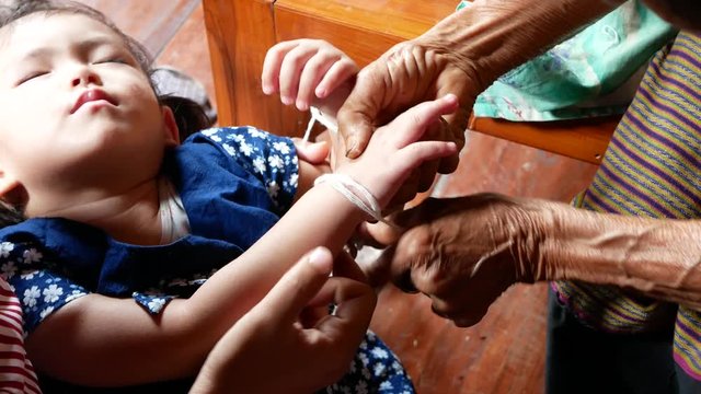 An elderly Thai person's hands tying holy white thread ( Sai sin ) around a sleeping baby's wrist at home - a gesture of goodwill / blessing ( Mad Mue ) from an elder 