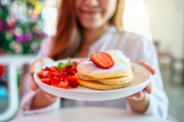 A woman holding and showing a plate of pancakes with strawberries and whipped cream