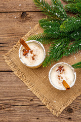 Eggnog cocktail in two glasses arranged with christmas decoration on old wooden table. Evergreen fer tree branch, artificial snow, sackcloth napkin
