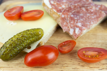 A slice of bread with salami and a slice of bread with cheese, while still small cocktail tomatoes and pickled cucumbers, served on a wooden board