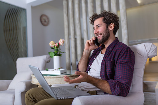 Young Attractive Smiling Guy Is Browsing At His Laptop And Talking Oh Mobile Phone, Sitting At Home On The Cozy Sofa, Wearing Casual Outfit. Freelance Business Work From Home Concept