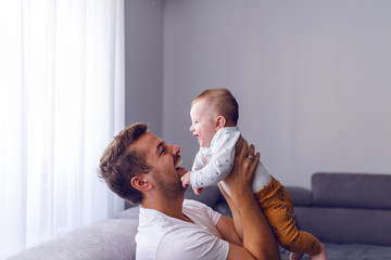 Playful handsome caucasian young dad lifting his loving 6 months old baby boy while sitting on sofa in living room. Baby enjoying time with father and laughing. - obrazy, fototapety, plakaty