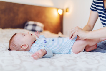 Adorable baby boy lying on bed while his mother changing his clothes before bedtime. Bedroom interior.