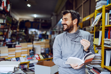Handsome caucasian smiling man standing in bookstore with book in hands and looking away.