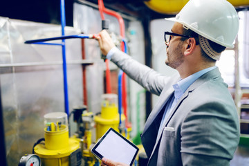 Handsome caucasian supervisor in suit and with helmet on head holding tablet and checking on boiler...