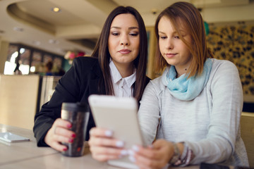 Two young women female friends using digital tablet at the restaurant cafe sitting by the table reading messages or browsing internet watching video or working shopping online