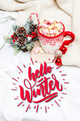 Red mug with marshmallows and winter ornaments on a white sheets with text Hello Winter