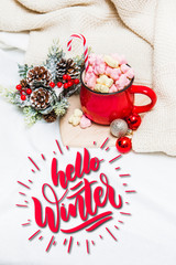 Red mug with marshmallows and winter ornaments on a white sheets with text Hello Winter