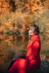 A girl in a red cloak sits on a stone in an autumn park near the lake
