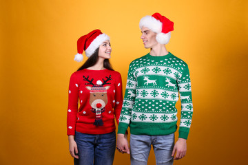 Studio portrait of young couple, boyfriend & girlfriend wearing santa claus hat & ugly christmas sweater. Holiday outfit w/ snowflake pattern print. Close up, copy space for text, isolated background.