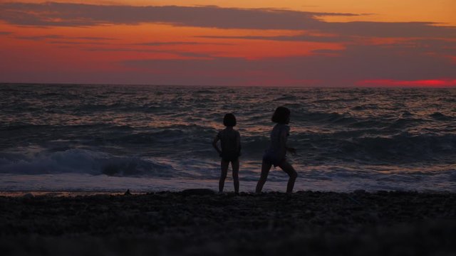Two little girls stand by the sea or ocean at sunset. Two childrens silhouettes at the shore. Waves splashing. Beautiful nature landscape.