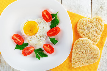 White plate with cherry tomatoes, parsley, fried egg in the shape of a heart and bread on a yellow napkin. Romantic food.