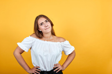 Confused young woman keeping hand on waist, looking disappointed isolated on orange background in studio in casual white shirt. People sincere emotions, lifestyle concept.