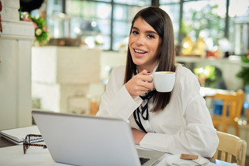 Young attractive caucaisan businesswoman in shirt sitting in cafe and enjoying coffee at break. On table is laptop.