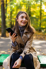 Young beautiful woman in a coat writes a message in the phone while sitting on a bench in the autumn park