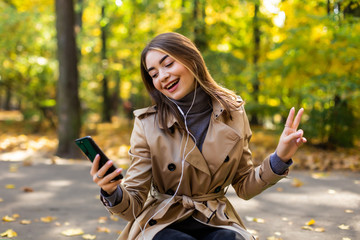 Young beautiful woman in a coat writes a message in the phone while sitting on a bench in the autumn park