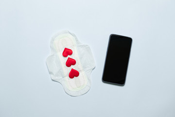 Menstruation period concept of slim cotton menstruation pad with red love shape as blood drops, smartphone on blue background.