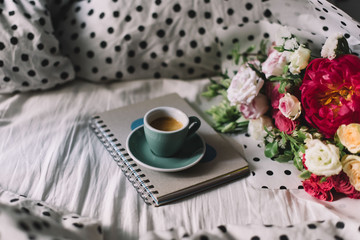 Delicious fresh morning cup of espresso coffee standing on the paper notebook in bed with beautiful tender flower bouquet of blossoming peony, eustoma, roses