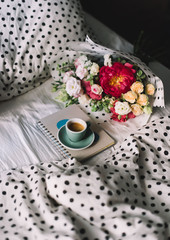 Delicious fresh morning cup of espresso coffee standing on the paper notebook in bed with beautiful tender flower bouquet of blossoming peony, eustoma, roses